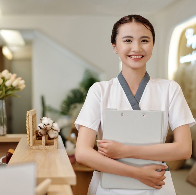 asian woman carrying notes stands in front of a desk in a spa or salon ready to welcome guests